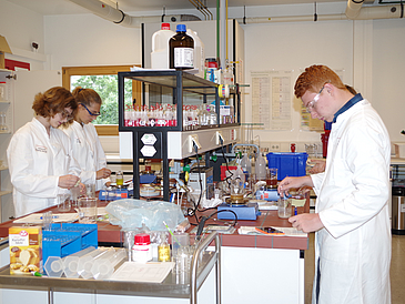 Junge Frauen und Männner stehen in einem Labor mit Schutzbrille und untersuchen Lebensmittel.